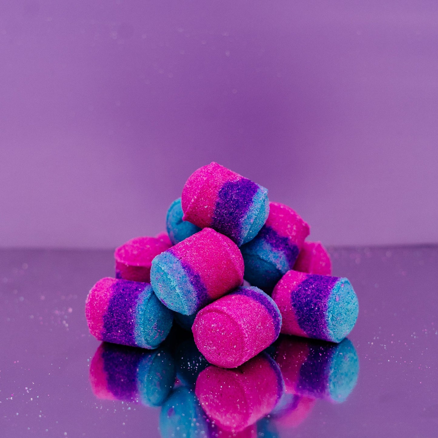 Birthday Cake Mini Bath Bombs Stack of pink and blue striped candies on a purple reflective surface with a purple background by Stellia Beauty