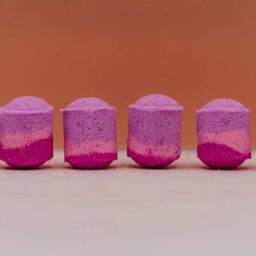 Four fairy dust purple and pink bath bombs on a beige surface with a brown background by Stellia Beauty
