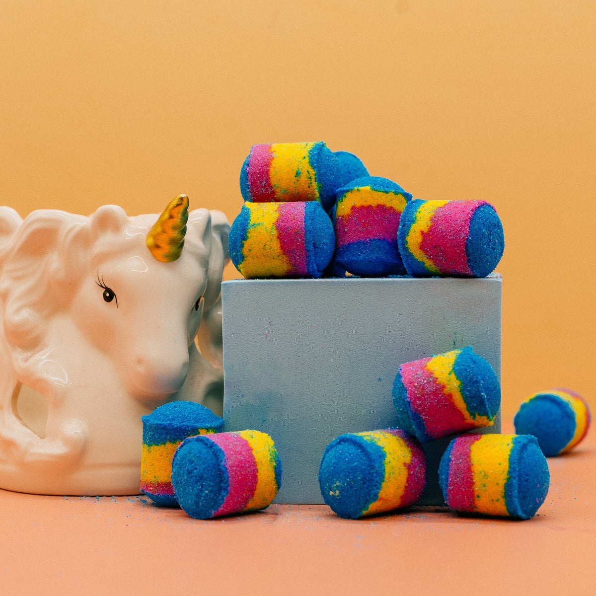 Unicorn Sparkle Shot Bombs Colourful striped bath bombs with a unicorn figurine on an orange background by Stellia Beauty